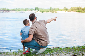 back view of father pointing on something at river to son at park
