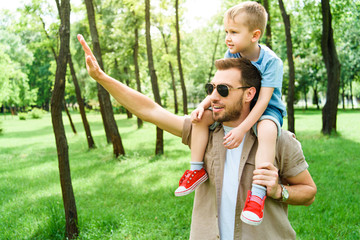 Obraz premium father waving hand to someone and holding son on shoulders at park
