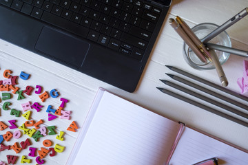 A keyboard, pencils, notebook and multicolor letters on the white wooden desktop.