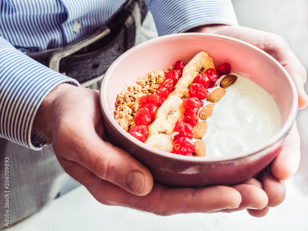 Wall mural Berry smoothie, muesli with nuts, dry berries, pieces of banana, beautiful, blooming flowers in a pink plate and men's hands. Concept of healthy and tasty food