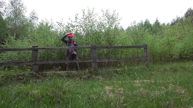 Mature bearded man traveler is happy meeting friends in woods. With backpack behind his shoulders goes along fence and the young forest
