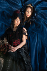 Young beautiful women in shiny blue dress standing behind women in black dress sitting at front, costume a fairytale, fantasy story.