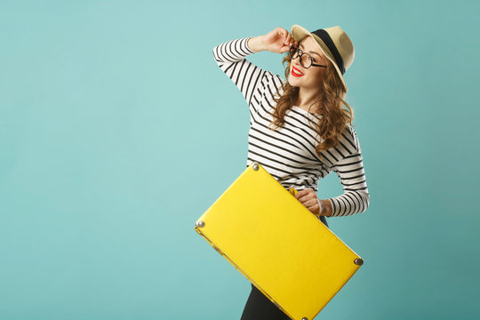 Young happy beautiful woman in hat and funny toy glasses holding yellow suitcase over blue background
