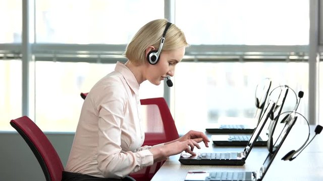 Woman with headset working in call center. Caucasian blond mature lady in office typing and smiling, side view, huge bright windows background.