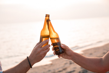 Cheers! Close up of hands toasting with bottles of beer in the beach. Celebration concept