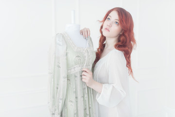 Tender portrait of a young dreamy redhead woman in nightdress. She is standing in dress room and planning to wear beautiful vintage dress.