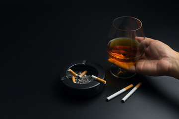 hand holding a glass of brandy, cigarettes and black ceramic ashtray full of ashes with the...