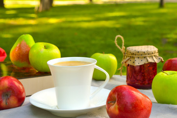 Cup of coffee in the garden on the table with green apples and red apples and jar of berries jam