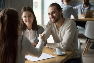 Happy millennial couple handshaking trusted broker signing mortgage contract, satisfied customers and realtor shaking hands making real estate deal, buying insurance or services at meeting in cafe