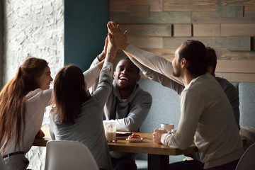 Diverse excited best friends raising hands giving high five together at meeting in cafe, happy...