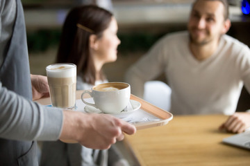 Waitress holding tray with cappuccino and latte, female hands serving hot aromatic brewed coffee drinks to couple visitors waiting at cafe table, coffee house shop service concept, close up view