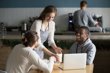 Smiling friendly waitress serving coffee drinks to diverse male friends meeting at cafe table, cafeteria server and multiracial visitors laughing together at funny joke in modern coffee shop house