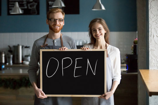 Smiling waiter and waitress holding chalkboard with open sign, cafe or coffee shop house business owners partners looking at camera welcoming inviting advertising new cafeteria bistro bar, portrait