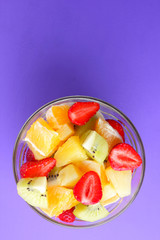 Fruit salad from pineapple, kiwi, orange, strawberry. Multicolored sliced fruit in a transparent dish. Vegetarian food on a violet background. Copy space