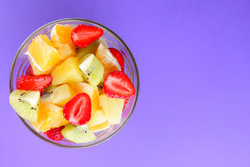 Fruit salad from pineapple, kiwi, orange, strawberry. Multicolored sliced fruit in a transparent dish. Vegetarian food on a violet background. Copy space
