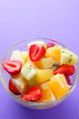 Fruit salad from pineapple, kiwi, orange, strawberry. Multicolored sliced fruit in a transparent dish. Vegetarian food on a violet background. Copy space