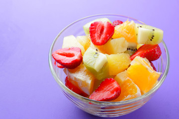 Fruit salad from pineapple, kiwi, orange, strawberry. Multicolored sliced fruit in a transparent dish. Vegetarian food on a violet background. Copy space