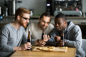 Excited diverse male friends having fun watching football game online video on smartphone drinking beer eating pizza together, three millennial multiracial men having fun cheering favorite sport team - Powered by Adobe