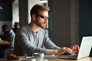 Focused millennial redhead man in glasses using laptop sitting at cafe table, serious businessman...