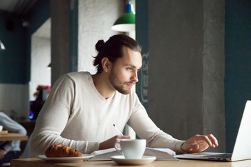 Focused man writing making notes studying with laptop in cafe, serious millennial student, businessman or entrepreneur using computer for e-learning and online education in public place concept