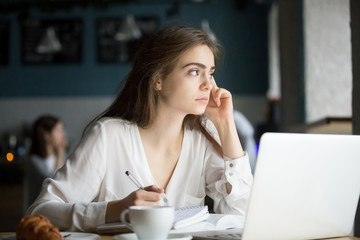 Thoughtful female student writing notes studying or working in coffee shop, serious pensive woman looking away searching for inspiration and new ideas sitting in in public place having writers block
