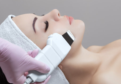 The doctor-cosmetologist makes the apparatus a procedure of ultrasound cleaning of the facial skin of a beautiful, young woman in a beauty salon. Cosmetology and professional skin care.