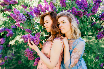 Adorable happy cheerful fabulous twin sisters in different beautiful summer dresses posing outdoor.  Similar cute female models in lilac colorful bushes with bloming flowers in park portrait.  Family.