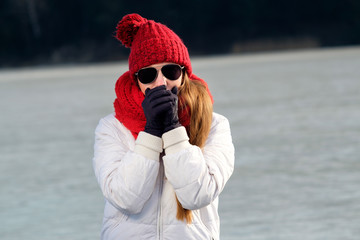 girl in aviator sunglasses, red cap, red scarf and white winter jacket freezes in winter, hugging herself in an attempt to get warm