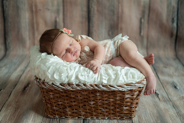 Newborn baby girl in bandage bow sleeping in wicker basket on plaid shawl on beige wooden floor