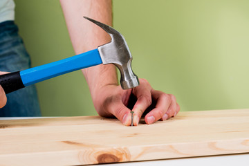 Hammering a nail into wooden plate. Concept of renovation, housework. The man is holding a blue hammer in his hand, holding a nail in the other hand. Handyman, DIY.