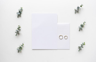 Wedding invitation card on white marble with wedding rings and oregano branches. Top view.