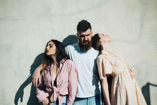 Bearded man standing between two beautiful women over white wall. Man making up his mind and choosing between impudent brunette and honest girl, love triangle, relationship problems concept