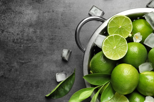 Colander with fresh ripe limes and ice cubes on gray background, top view