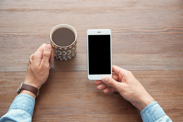 Young man holding mobile phone with blank screen and cup of coffee in hands at wooden table