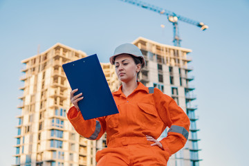 beatiful business woman engineer wearing orange coverall is standing with plan papers. Construction project with crane is on background