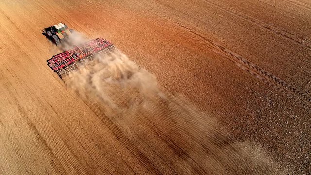 Big farm tractor tilling dusty Springtime fields, aerial view.
