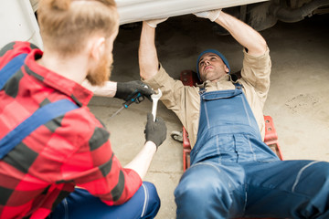 Fototapeta na wymiar Young professional auto technician lying under truck and fixing breakdown while his colleague consulting him and giving tools