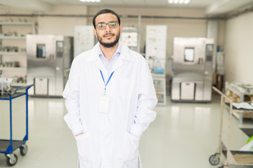 Serious confident handsome young Arabian factory lab technician in lab coat wearing badge and...
