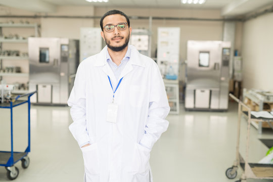 Serious Confident Handsome Young Arabian Factory Lab Technician In Lab Coat Wearing Badge And Eyeglasses Holding Hands In Pockets And Looking At Camera In Modern Innovation Plant.