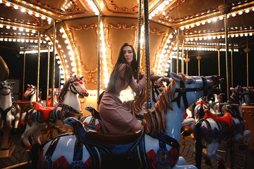 Young beauty model woman posing with old horse carousel in summer park with magic lights