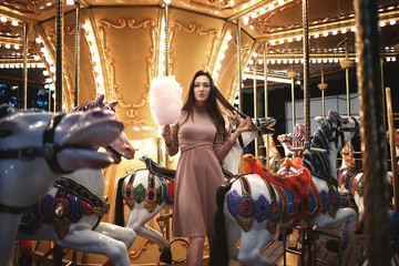 Plakat Young beauty model woman posing with old horse carousel in summer park with magic lights