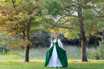 full length portrait of blonde girl wearing greensand white medieval costume, standing pose. walking through a forest.