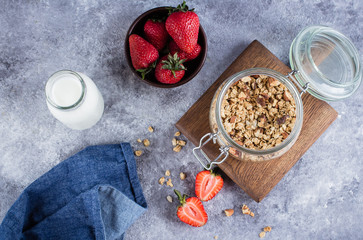 Healthy breakfast ingredients. Homemade granola in open glass jar, milk or yogurt bottle, strawberry on gray concrete background, top view, copy space