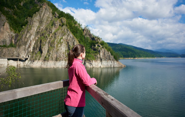 Fototapeta premium Brunette female tourist looking far into the distance admiring magnificent views of green rocky mountains and breathtaking Vidraru lake in Romania. Woman amazing beautiful scenery landscape happy calm