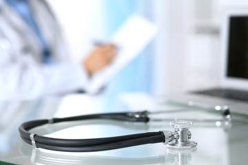 Stethoscope lying on glass desk with laptop computer at busy physician background. Medicine or pharmacy concept. Medical tools at doctor working table