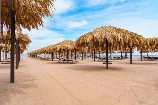 Beach straw umbrellas or palapas and sunbeds on the sandy deserted sea shore. Cargo ship is on far background. Cloudy blue sky.