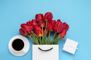 White Gift Bag, a small white gift box, a white cup with black coffee and a bouquet of red tulips on a blue background. Concept Offers an engagement or marriage, shopping