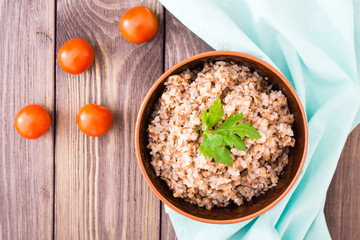 Cooked buckwheat in a bowl with a leaf of parsley and cherry tomatoes on a wooden table. Top view
