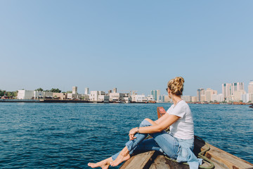 Fototapeta na wymiar Young beautiful blonde woman sitting at boat and looking at sea and the city