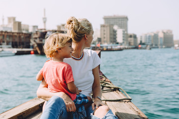 Young beautiful woman with her little son sitting at boat and contemplating the seascape of Dubai